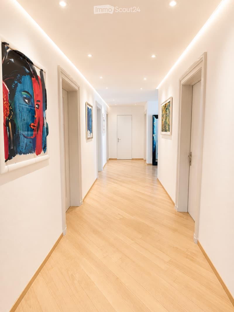 Bright, modern hallway with light wooden flooring, white walls, recessed lighting, and several doors leading to other rooms. Artwork is displayed on the walls.