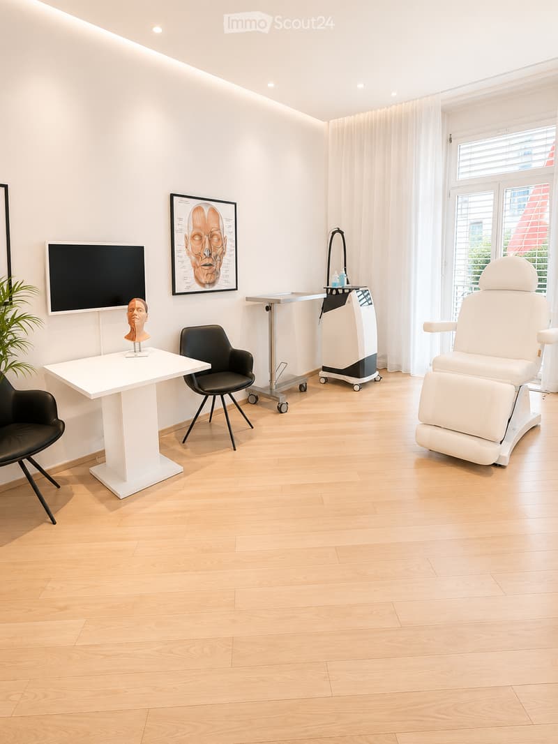 Modern, well-lit room with light wooden flooring, white walls, and large windows with curtains. Features a white table with a mannequin head, two black chairs, a large screen, a medical examination chair, and a small cart with medical equipment.