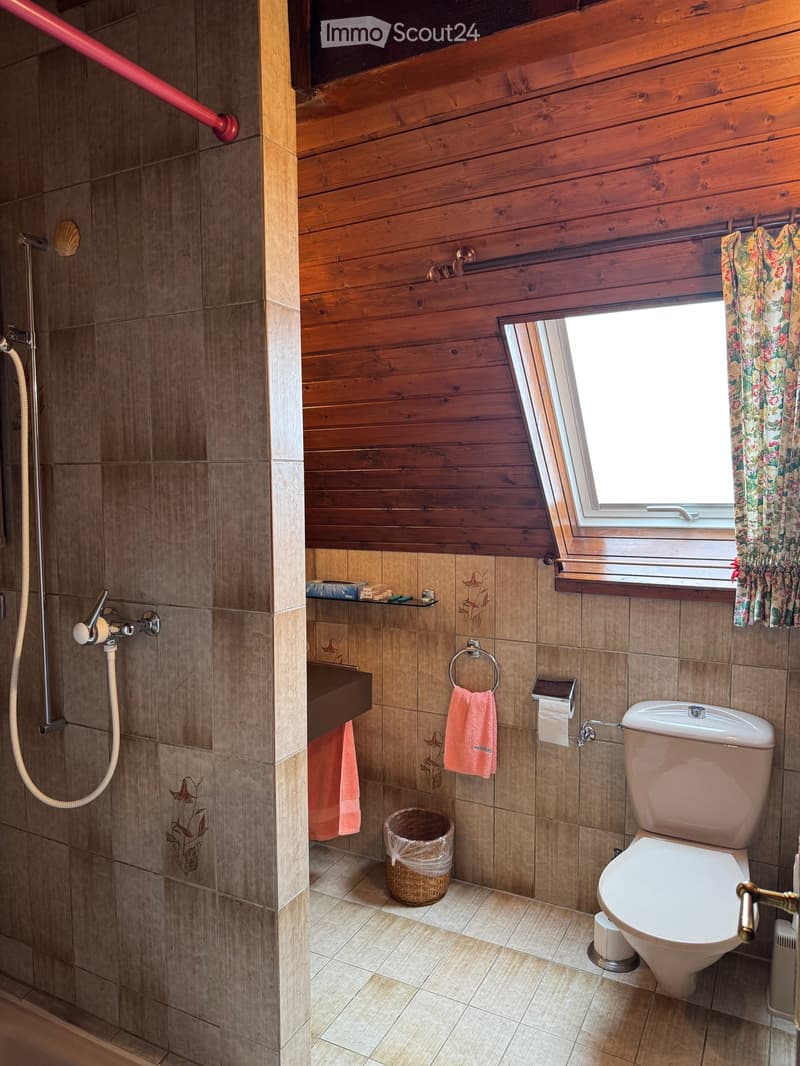 Bathroom with wooden walls, tiled shower area, toilet, sink, and window with floral curtain.