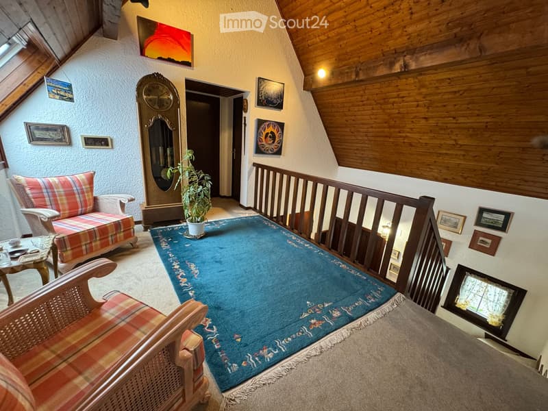 A cozy living room with wooden beams, a blue patterned rug, a plaid sofa, a wooden chair, a small table, a large antique clock, and several framed pictures on the walls.