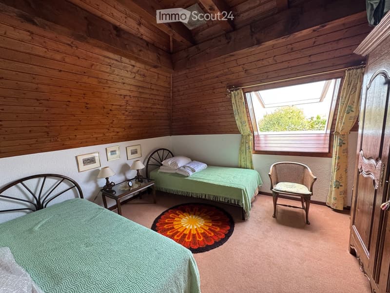 Cozy bedroom with wooden walls and ceiling, two single beds, a wicker chair, a small table with lamps, and a colorful round rug.