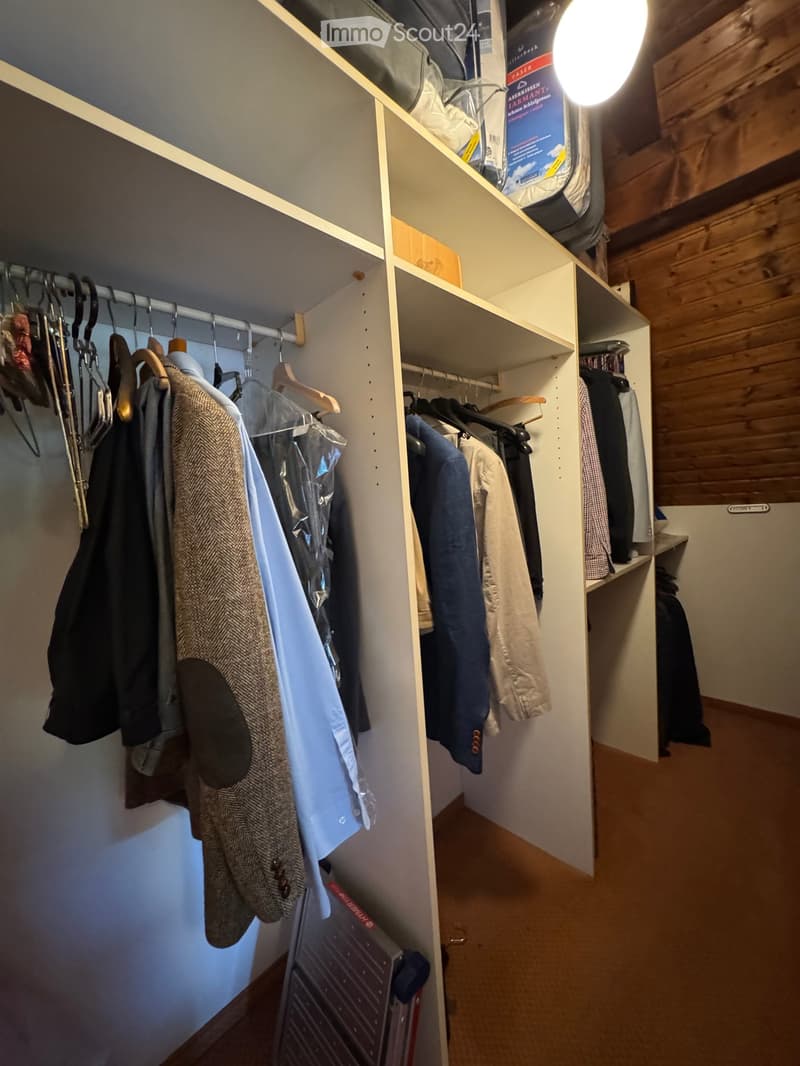 Well-lit walk-in closet with wooden shelves and hanging rods, containing various clothing items and storage boxes.