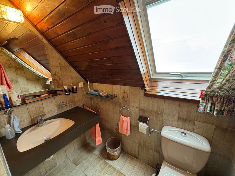 Bathroom with wooden slanted ceiling, tiled walls, sink, toilet, mirror, and various toiletries.