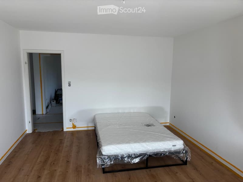 A bedroom with a wooden floor, white walls, and a single bed with a mattress and a protective cover. The room has a doorway leading to another room.
