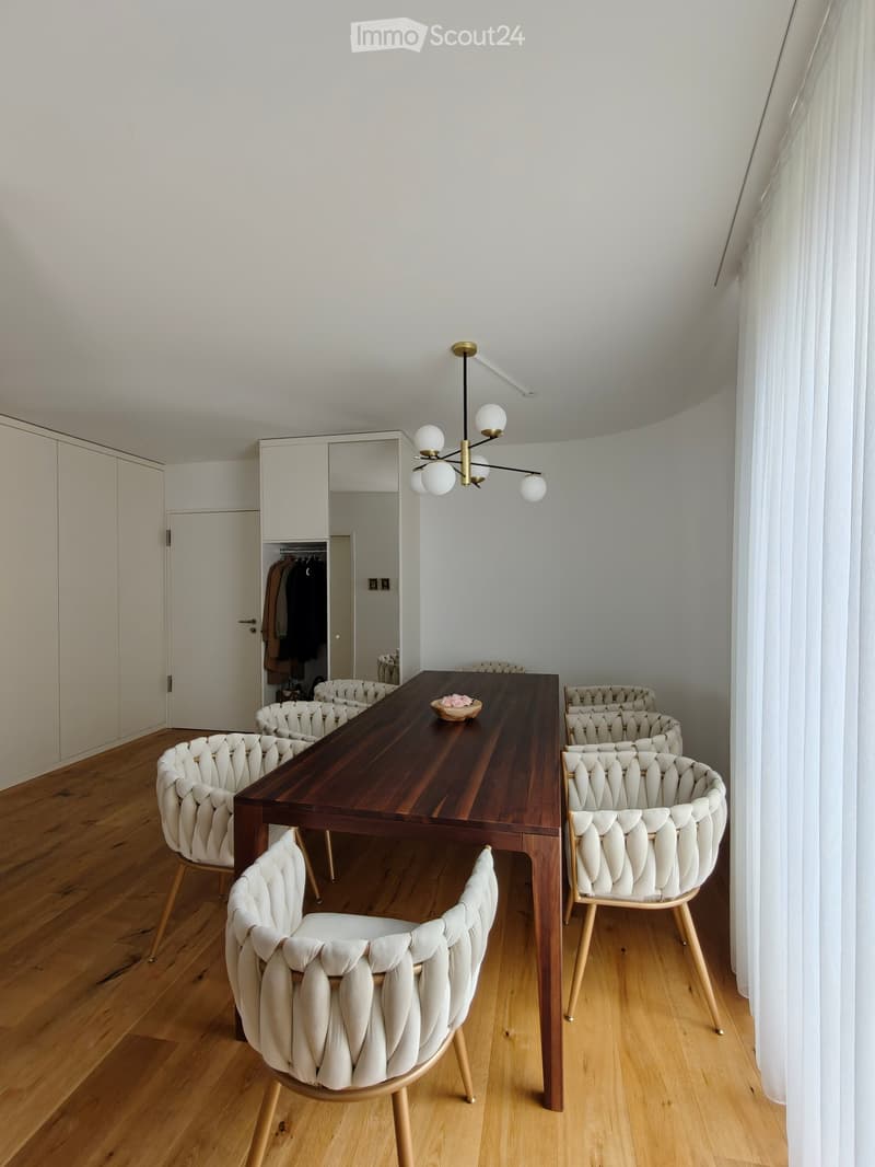 Modern dining area with a long wooden table, six white upholstered chairs, a chandelier, and a large mirror on the wall.
