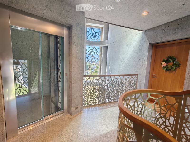 Modern hallway with concrete walls, glass elevator, decorative railing, and a wooden door with a floral wreath.