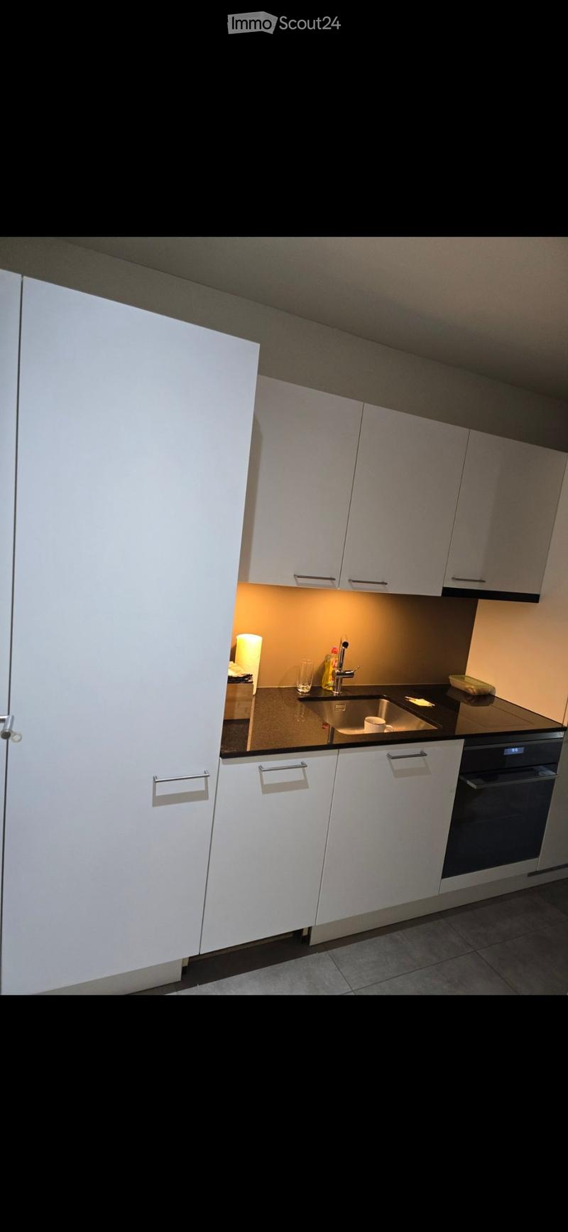 Modern kitchen with white cabinets, black countertops, and stainless steel appliances including a sink and oven. A glass and a bottle are placed on the counter.