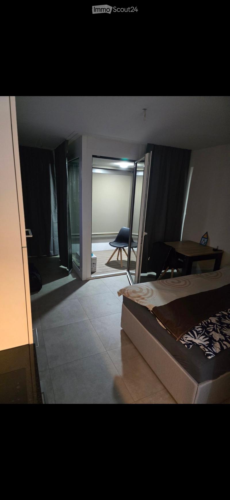 A small, modern bedroom with a bed, a chair, a small table, and a curtained window. The room has tiled flooring and an open door leading to a balcony area.