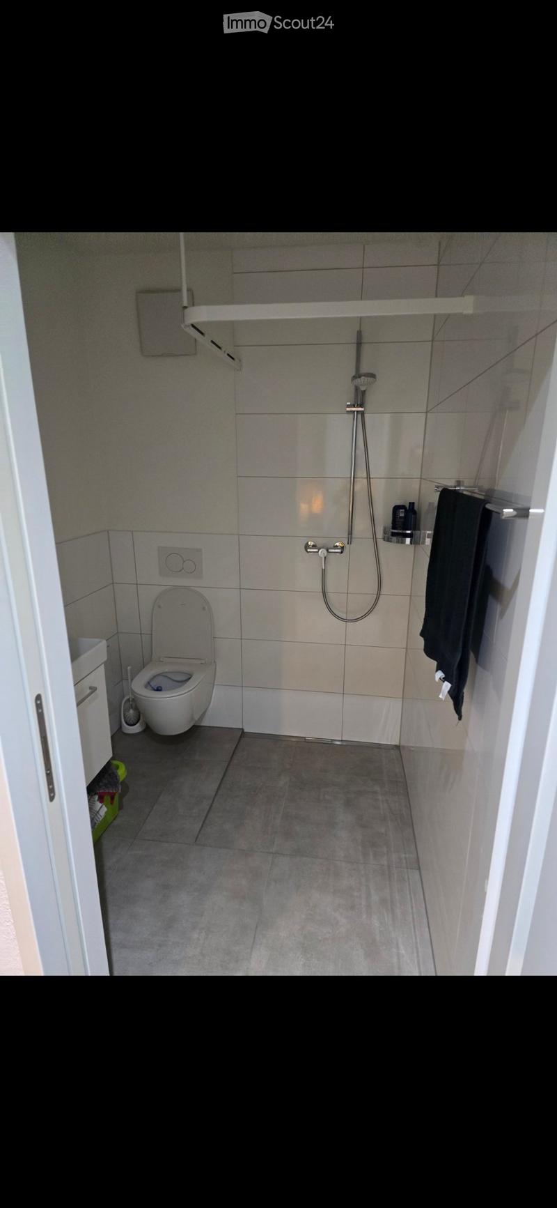 Modern bathroom with a toilet, shower, towel, and shower shelf. The walls are tiled in a light gray color, and the floor is covered with dark gray tiles.