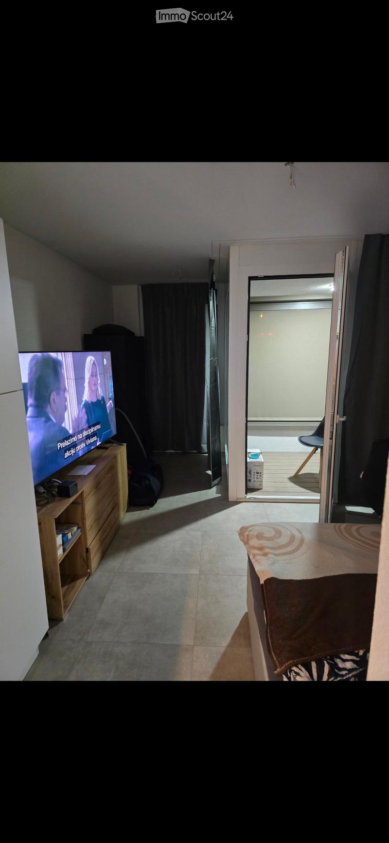 A furnished room with a bed, television on a wooden cabinet, and a chair near the balcony door.