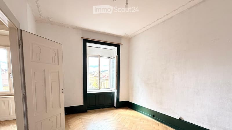 Empty room with wooden flooring, white walls, and a large window with a view of the cityscape. The room features a white door on the left and a green baseboard along the walls.