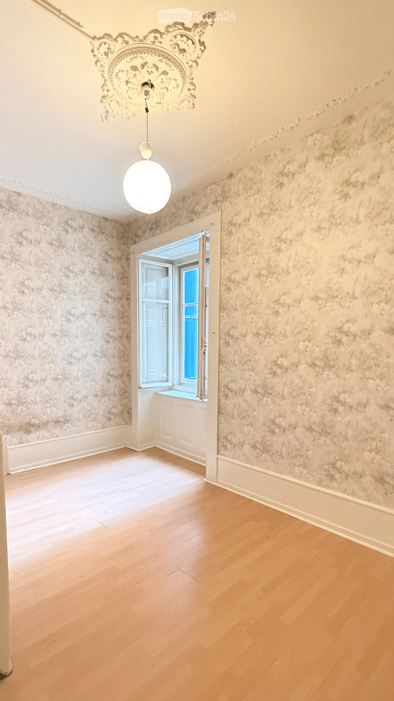 Empty room with light wooden flooring, textured walls, and a decorative ceiling light fixture. An open doorway leads to a windowed area.