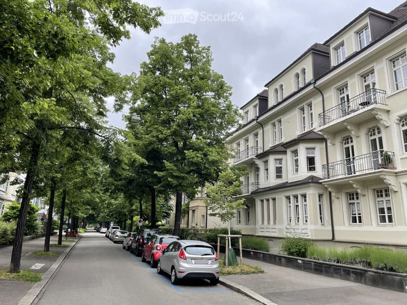 A multi-story residential building with balconies and large windows, situated on a tree-lined street with parked cars.