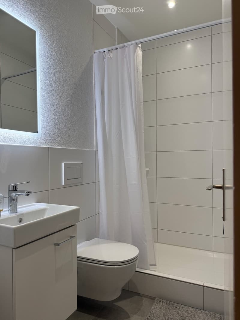 Modern bathroom with white tiles, a sink with a chrome faucet, a toilet, a bathtub with a white shower curtain, and a mirror above the sink.