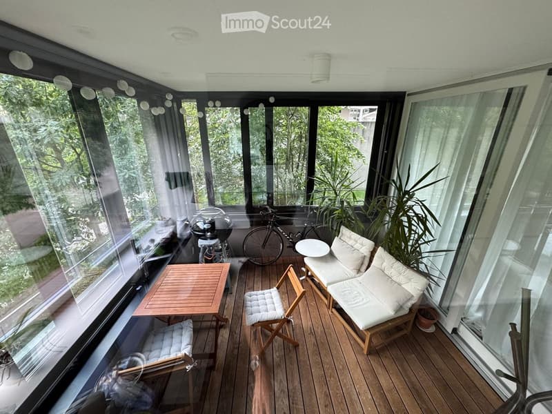 A modern, enclosed balcony with large glass windows, wooden flooring, and a cozy seating area including a table, chairs, and a sofa. A bicycle is parked near the window, and plants add a touch of greenery.
