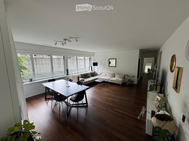 Bright living room with large windows, wooden flooring, a white sectional sofa, a dining table with chairs, and decorative mirrors on the walls.
