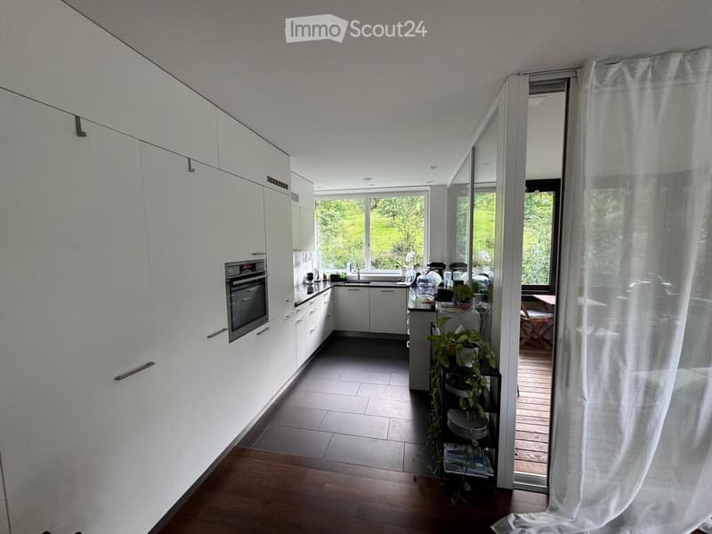 Modern kitchen with white cabinets, stainless steel oven, and large windows overlooking a green landscape. Includes a small seating area with a table and chairs, and a plant near the window.