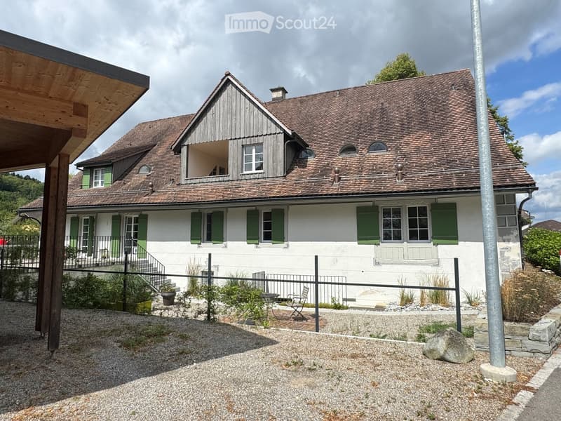 Two-story house with white walls, green shutters, and a tiled roof. Features a small balcony, a fenced garden area with a table and chairs, and a stone pathway.