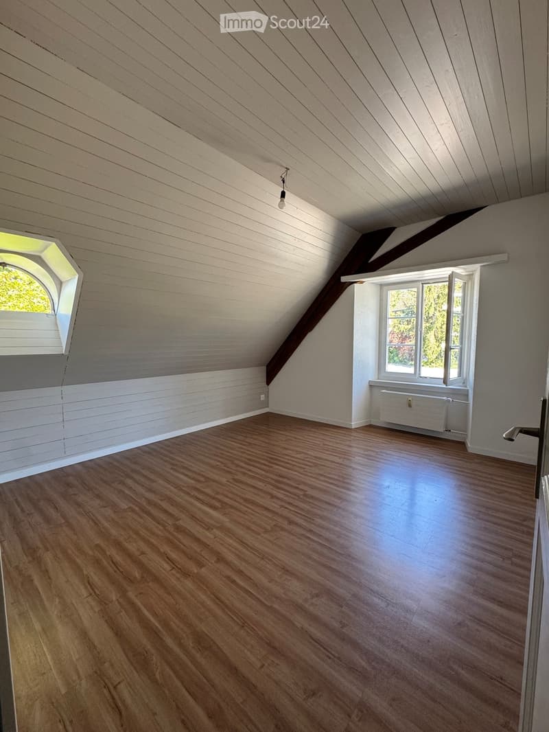 Empty attic room with wooden flooring, slanted wooden ceiling, and two windows, one of which is octagonal.