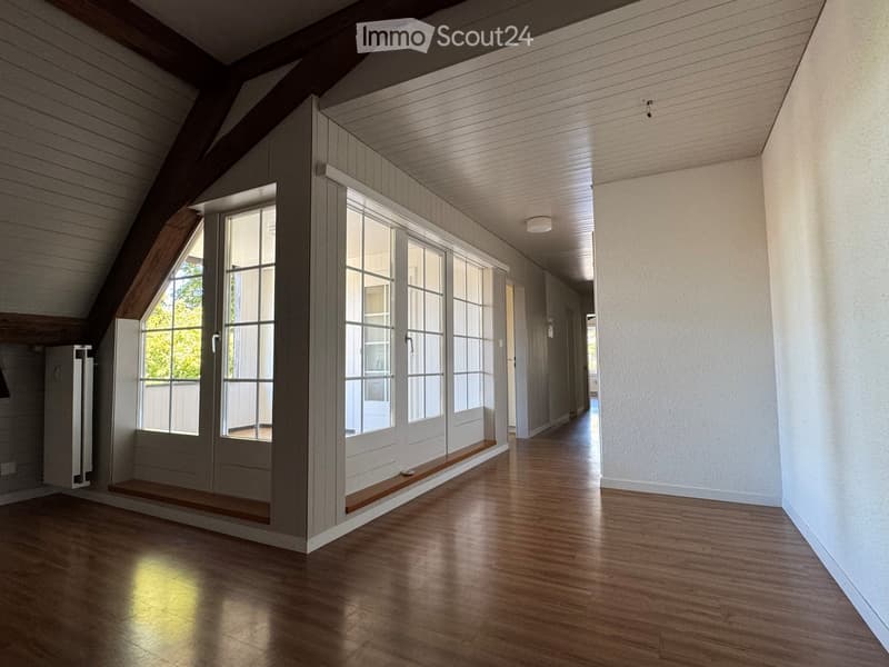 Empty attic corridor with wooden floors, white walls, large windows, and exposed wooden beams on the ceiling.