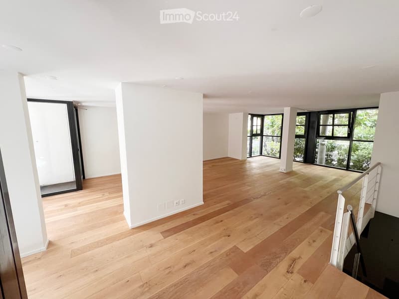Empty room with light wooden flooring, white walls, large windows, and a staircase leading upwards.