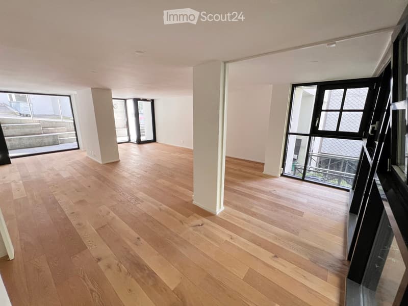Empty room with light wooden flooring, white walls, and large windows with black frames, leading to an outdoor area.