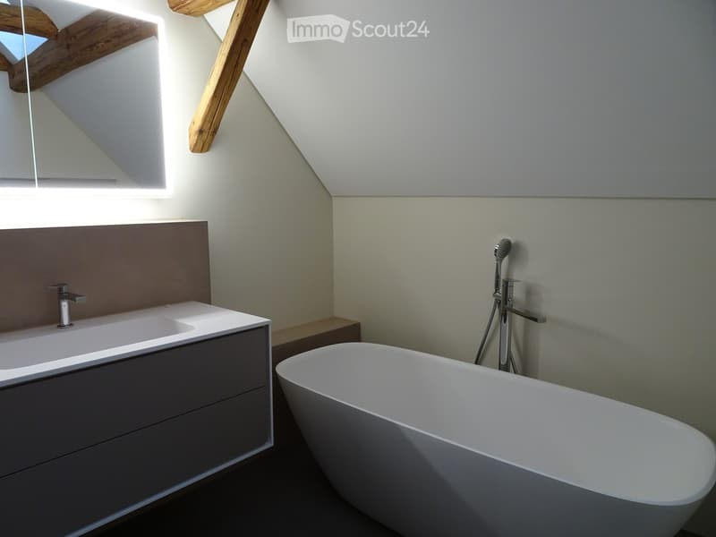 Modern bathroom with a freestanding bathtub, a sink with a sleek faucet, and a large mirror above the sink. The room features light-colored walls and wooden beams on the ceiling.