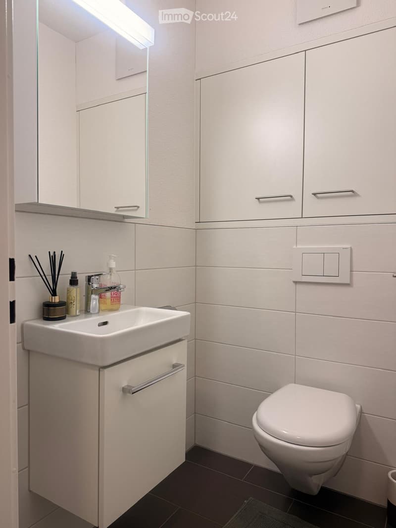 Modern bathroom with white tiles, a sink with a faucet, a toilet, a mirrored cabinet, and a light switch on the wall.