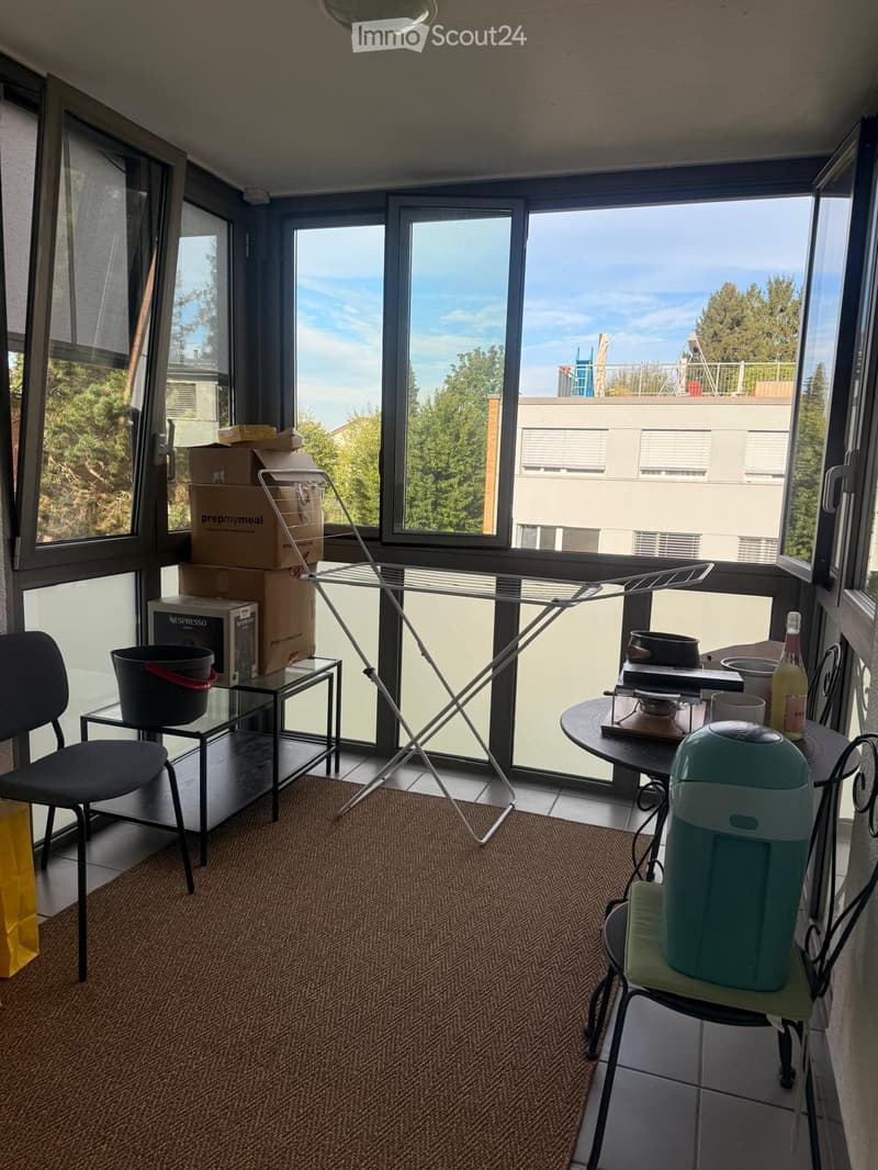 Balcony with large windows offering a view, equipped with a small table, chairs, a laundry rack, and some storage boxes.