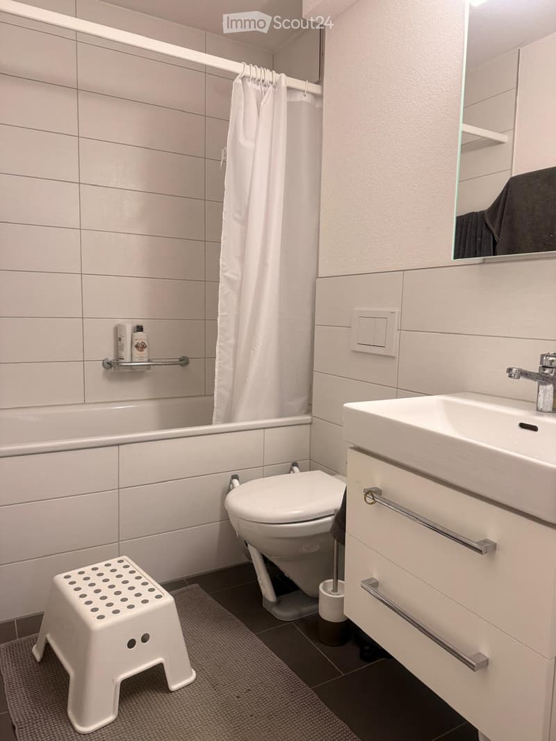 Modern bathroom with white tiles, bathtub, toilet, sink, mirror, and a small white stool. Includes a shower curtain and a bottle on the shelf.