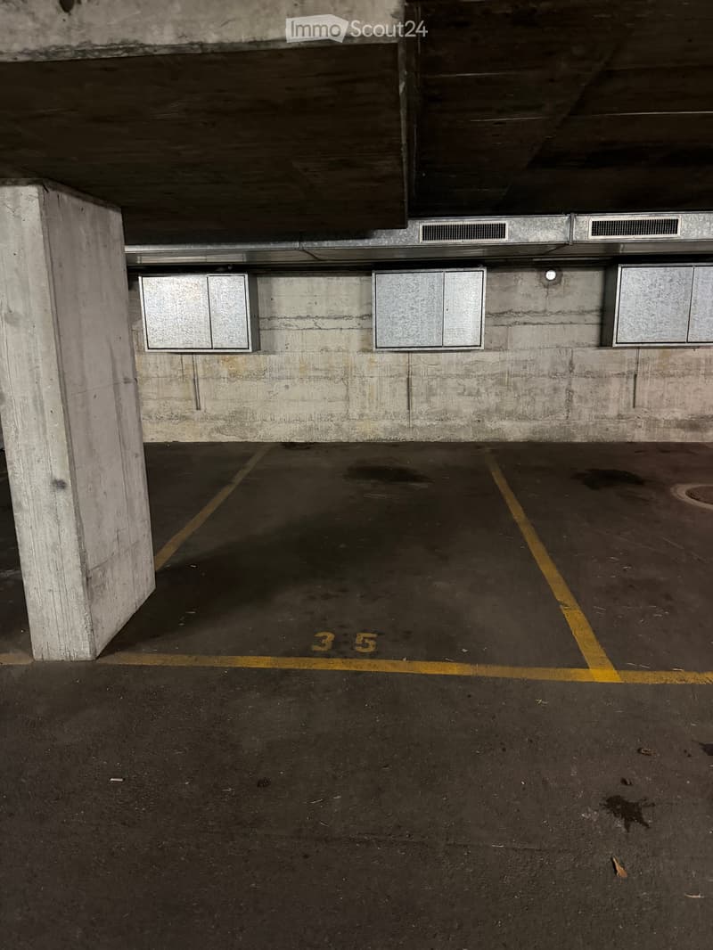 Underground parking area with concrete pillars, marked parking spaces, and ventilation grates on the ceiling.