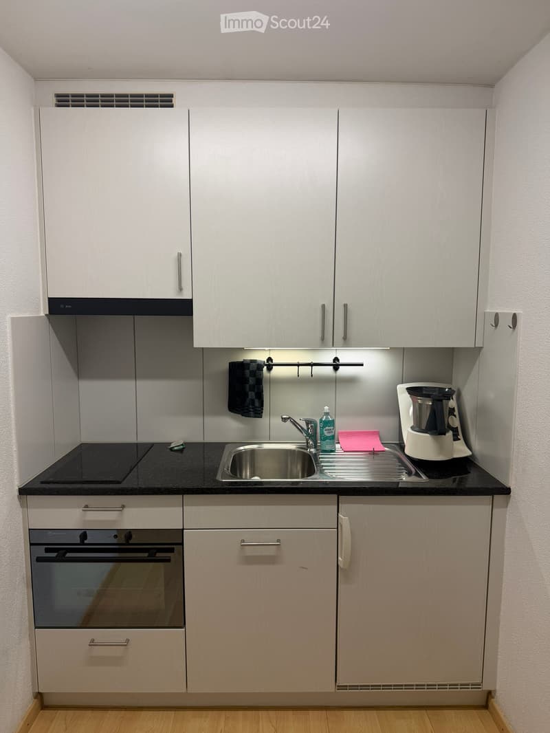 Modern kitchen with white cabinets, black countertop, stainless steel sink, and built-in oven. Coffee maker and cleaning supplies are on the counter.