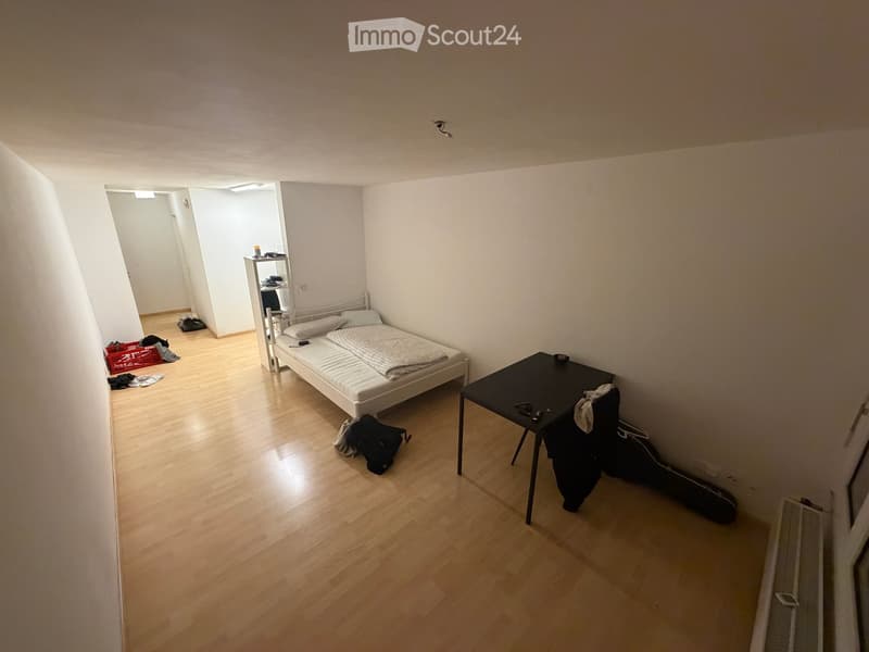 A small, furnished bedroom with a single bed, a small table, a chair, and a shelf in the corner. The room has wooden flooring and white walls.