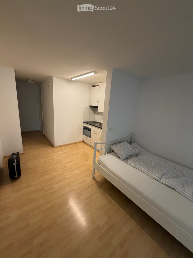 A small, furnished room with a single bed, a small kitchen area with cabinets and an oven, and a suitcase on the floor.