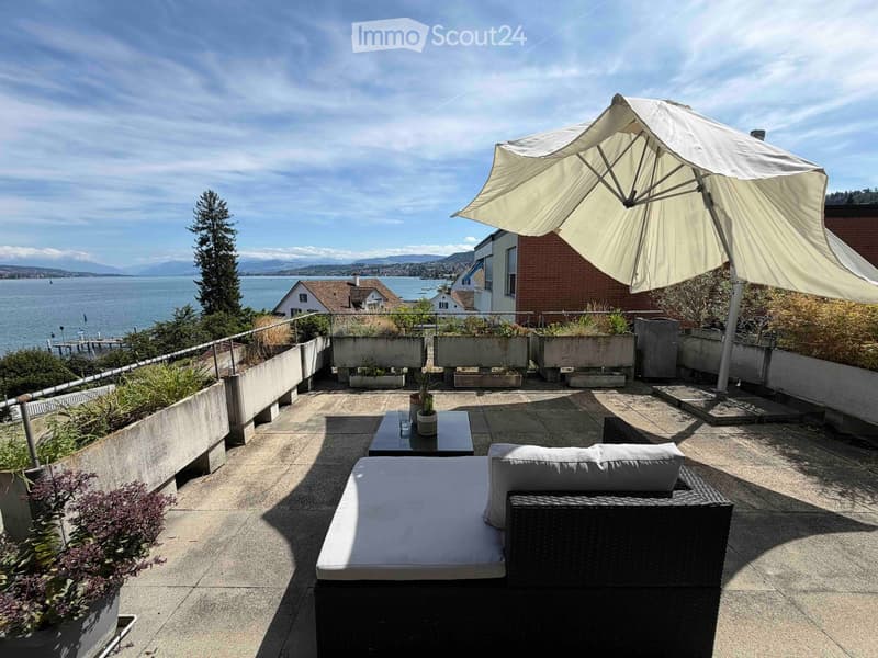 Balcony with a view of the lake and mountains, featuring a sofa, table, and umbrella.