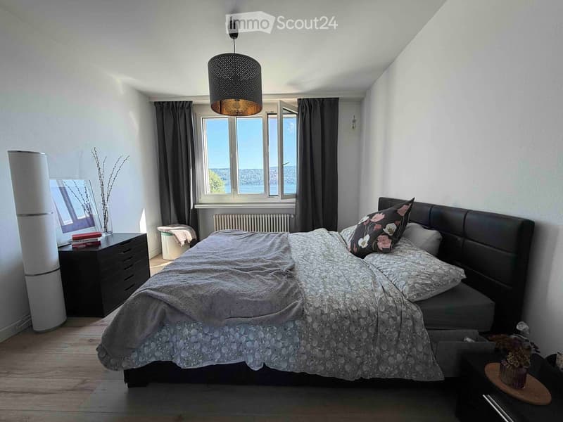 Modern bedroom with a large bed, nightstand, mirror, and a view of the city through large windows.