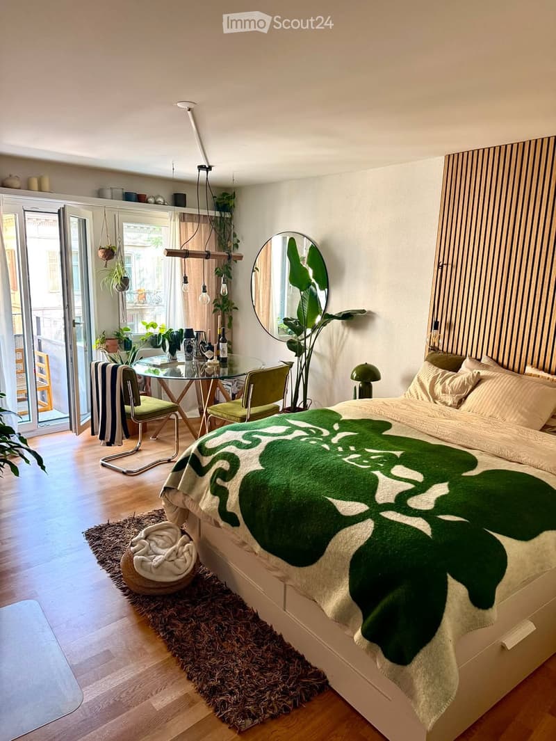 A modern bedroom with a large bed, green throw blanket, round mirror, hanging plants, and a small dining area with a glass table and chairs.