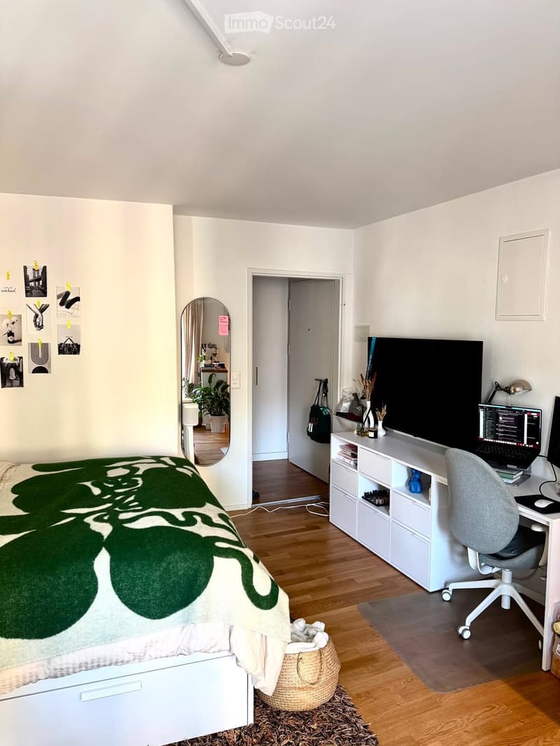 A well-lit bedroom with a bed, desk, chair, laptop, and a large screen. The room features a mirror, storage unit, and a small basket on the floor.