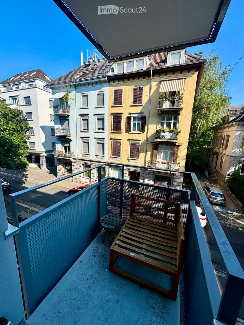 Balcony with a wooden bench and a small table, overlooking a street with multi-story residential buildings and parked cars.