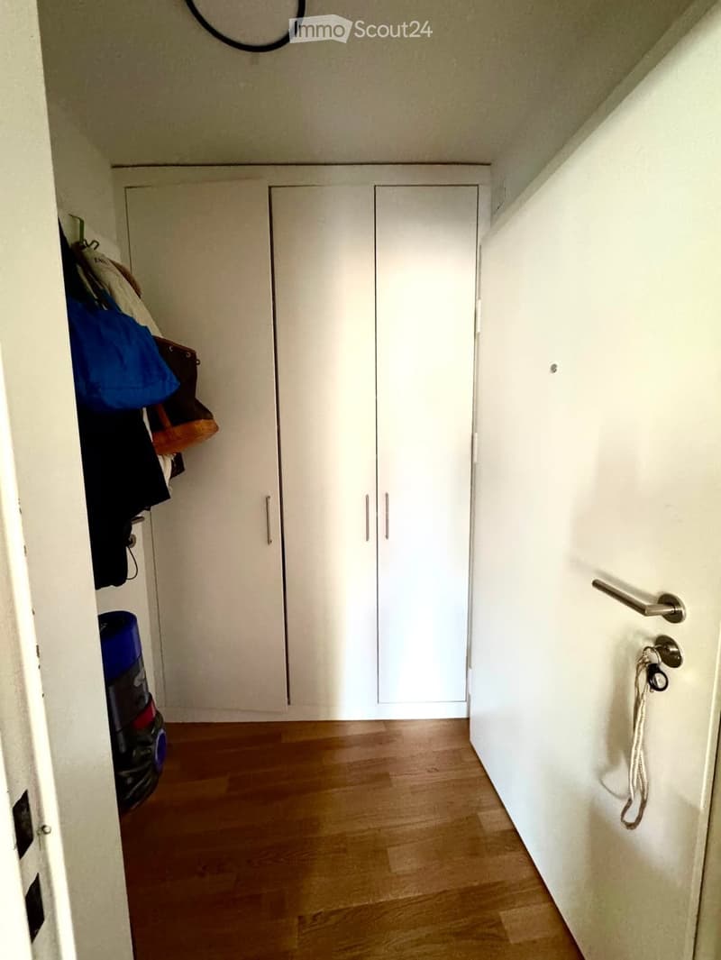 A narrow hallway with wooden flooring, white walls, and built-in white closet doors. A vacuum cleaner and some clothing are visible on the left side.