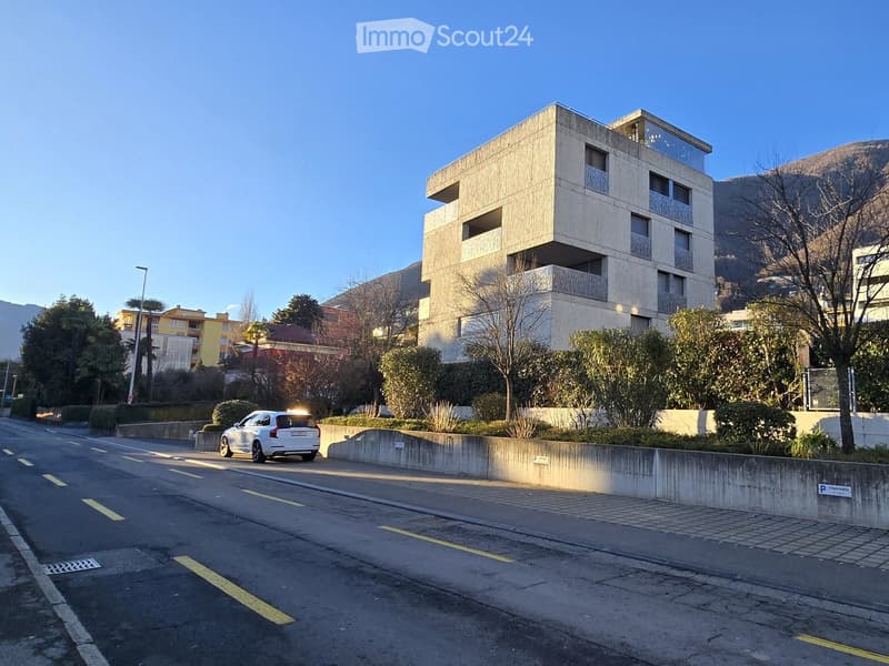 Modern building with a sloped driveway and balconies, surrounded by greenery and mountains
