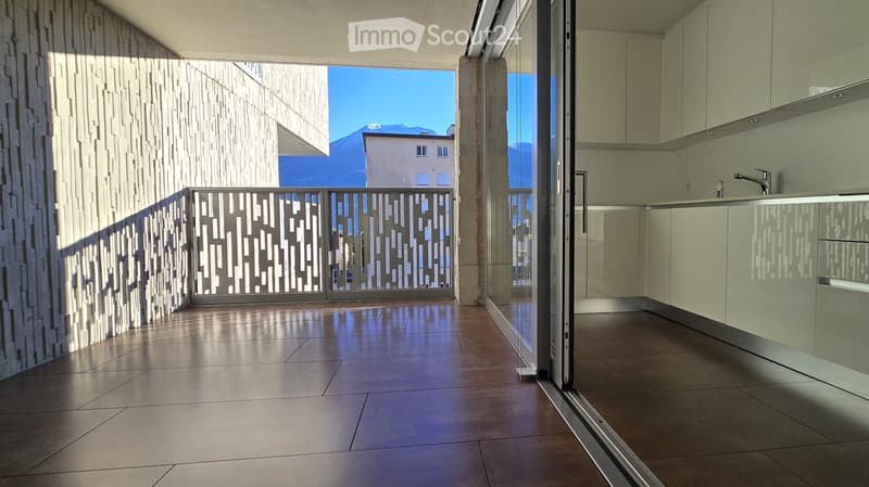 Balcony with railing, tiled floor, open to a kitchen area, large windows, distant mountains