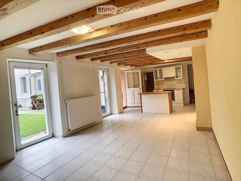 Bright, open-plan living and dining area with exposed wooden beams on the ceiling, large sliding glass doors leading to a garden, and a modern kitchen area with white cabinets and a central island.
