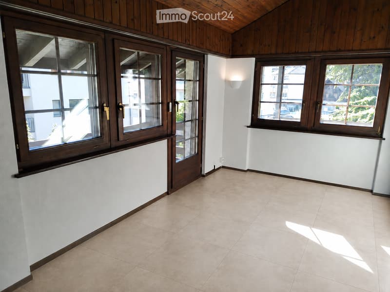 Empty room with tiled floor, white walls, large windows with wooden frames, and a wooden ceiling.