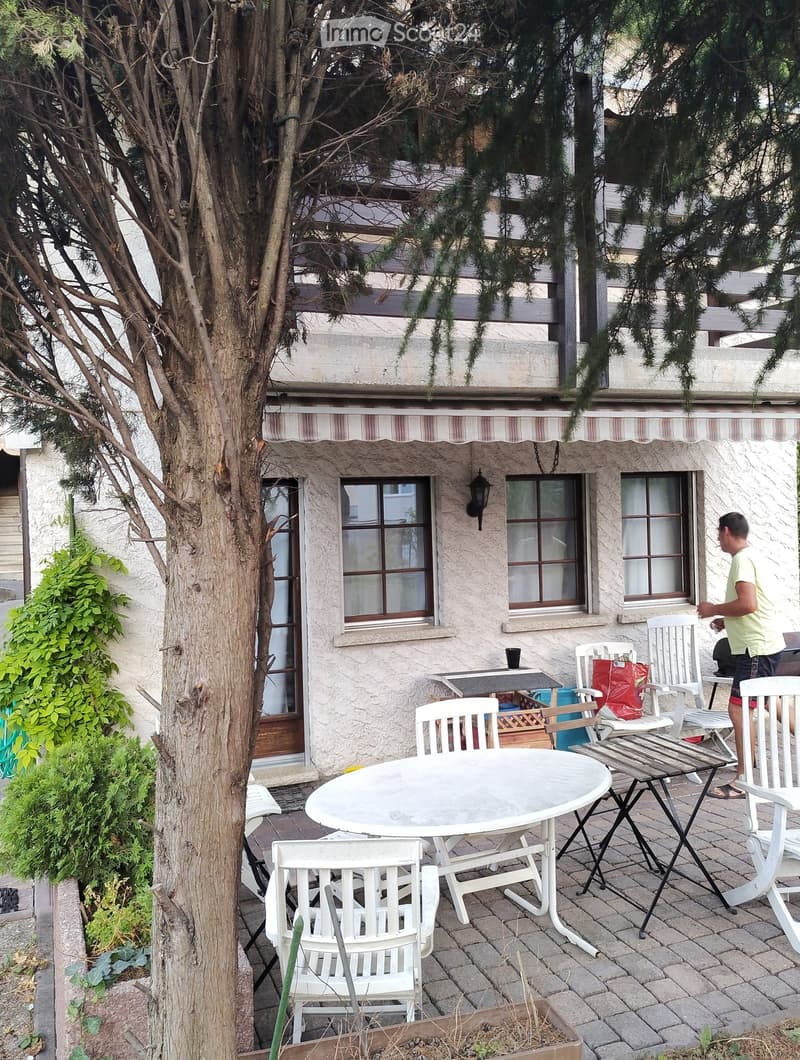 white stucco exterior, brown wood awning, large windows, balcony, front garden, patio with white chairs and tables