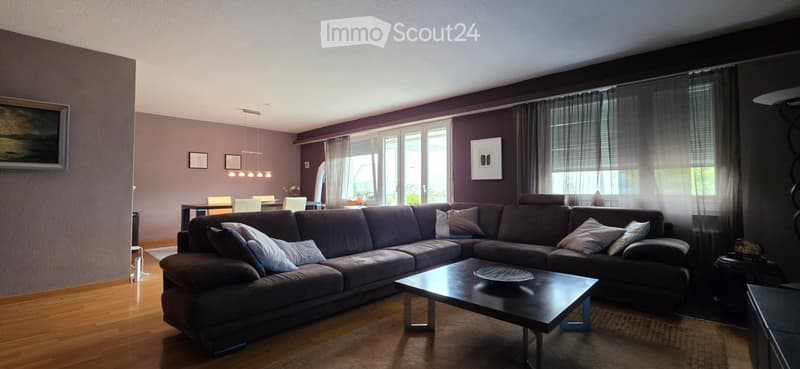 Modern living room with dark grey sectional sofa, coffee table, dining area with chairs, large windows with curtains, and artwork on walls.