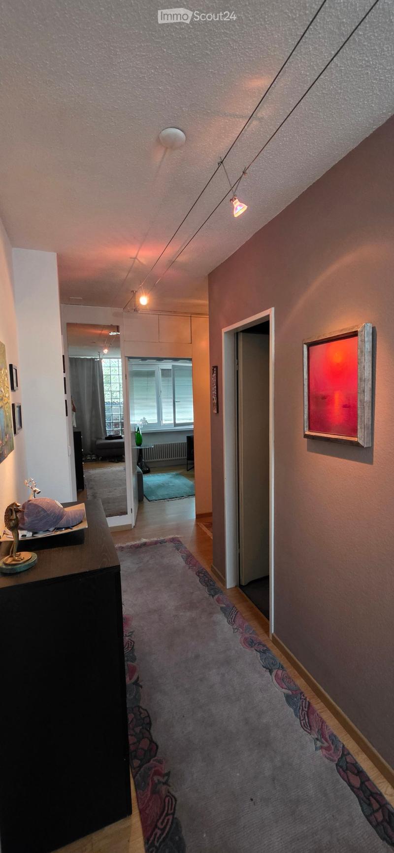 Hallway with wooden flooring, patterned rug, and warm lighting. Features open doors leading to other rooms, including a living area with a sofa and a dining area with a table and chairs.