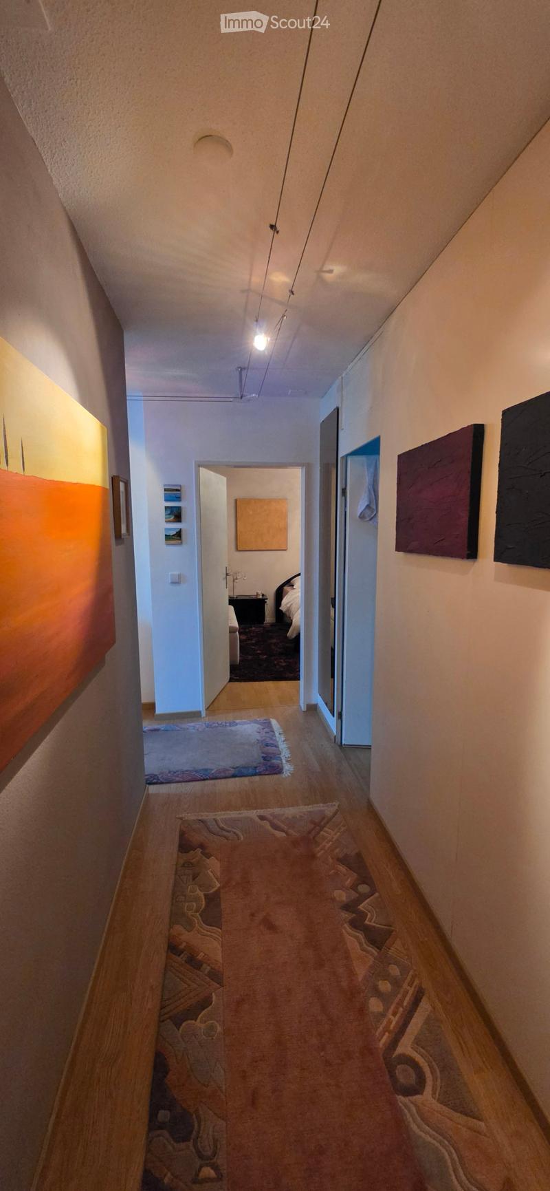 A narrow hallway with wooden flooring, warm lighting, and artwork on the walls. It leads to a bedroom with a bed and nightstand visible at the end.