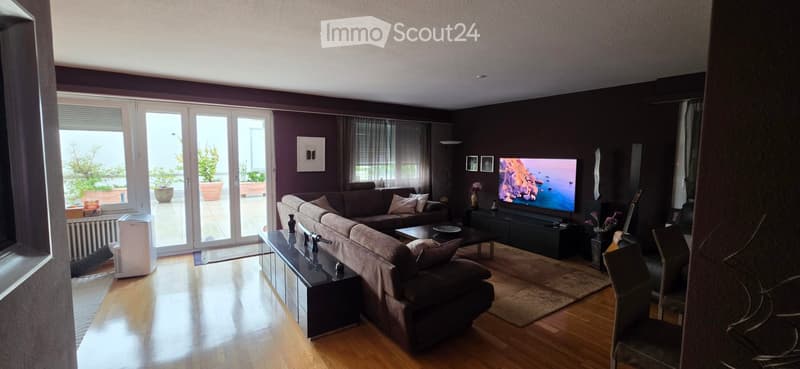 Modern living room with dark purple walls, large sectional sofa, coffee table, TV, and guitar in the corner. Large windows and sliding doors lead to a balcony with outdoor plants.