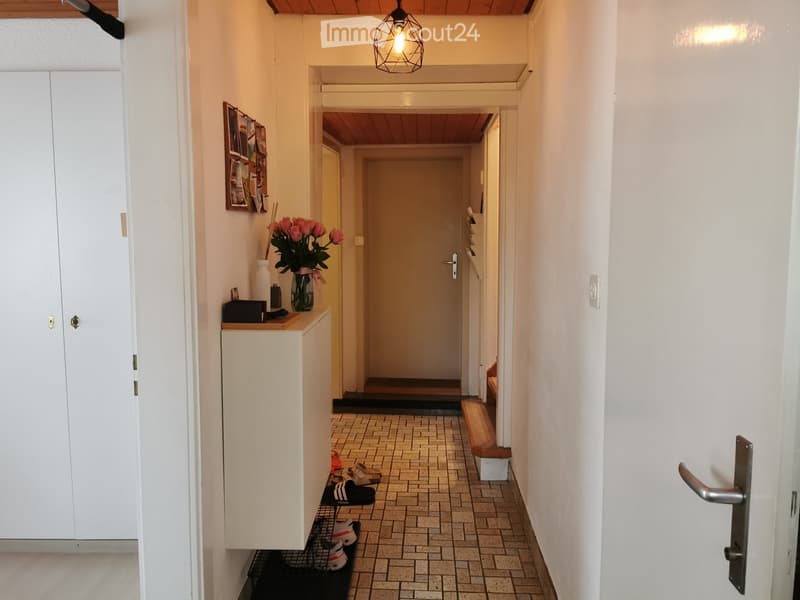 A narrow hallway with tiled flooring, white walls, and a small bench on the right side. A small table with a vase of flowers and a few items sits against the left wall, and several pairs of shoes are placed on a shoe rack near the entrance.